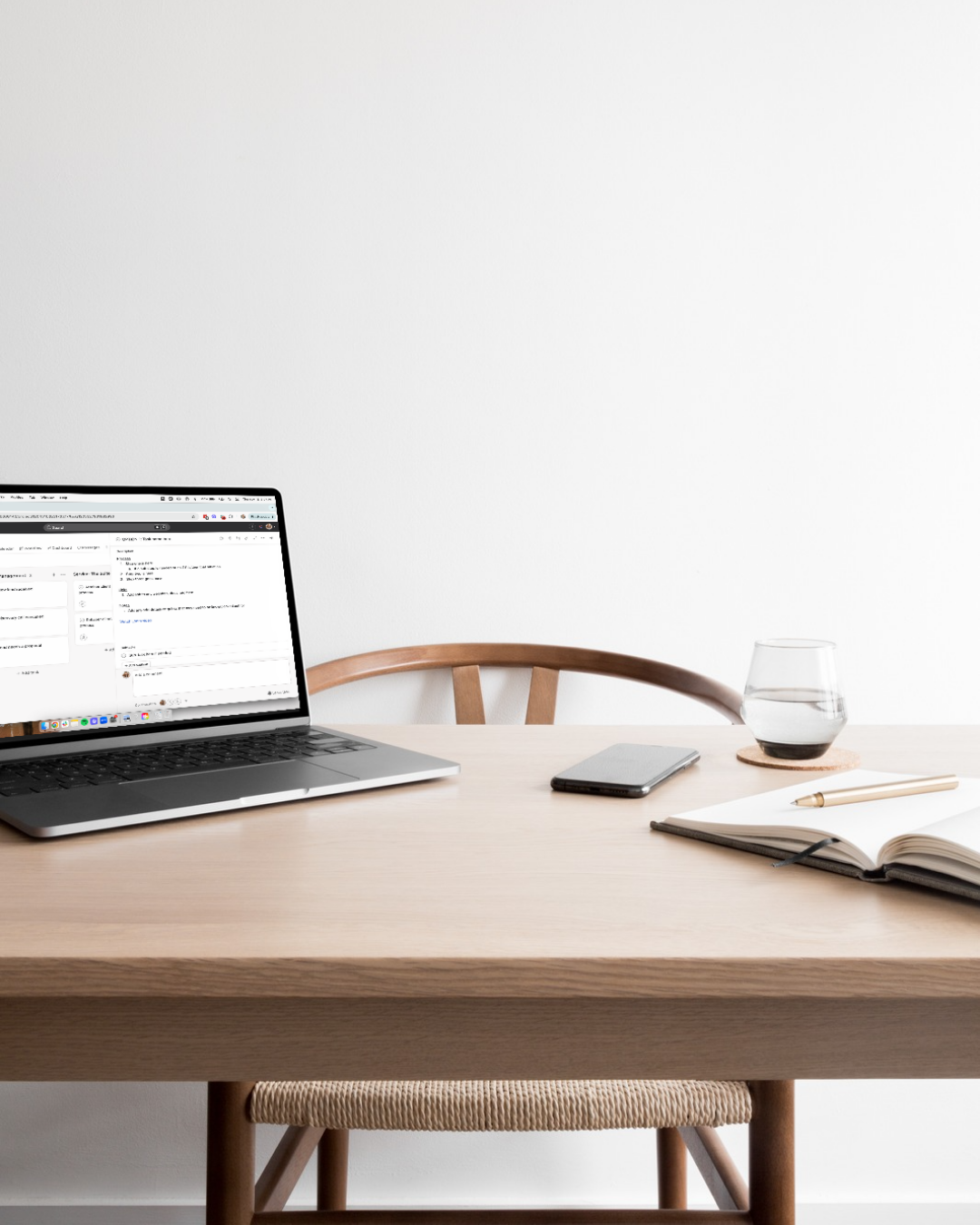 Laptop with Asana screenshot on table with phone glass of water and open journal